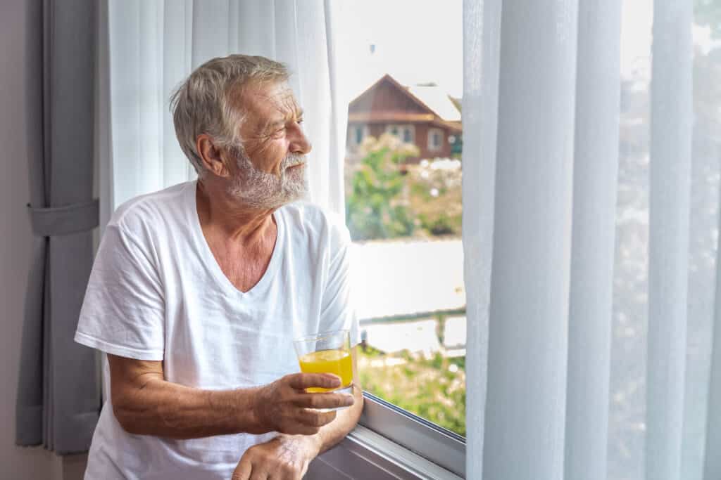 senior elderly man standing looking out window bedroom after waking up morning with juice