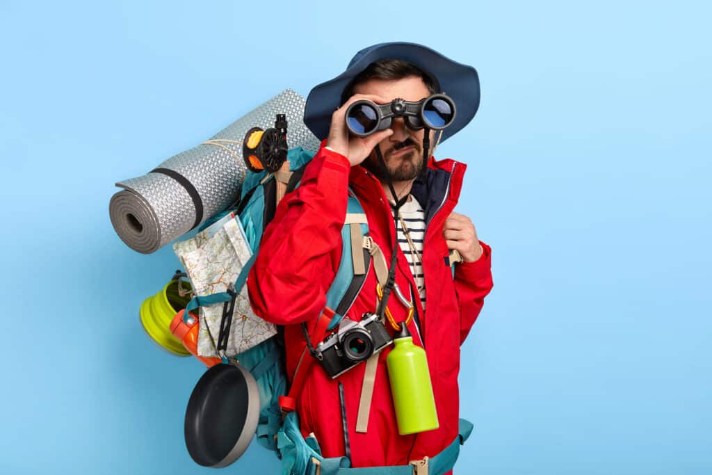 serious unshaven male backpacker keeps binoculars near eyes wears hat red jacket explores new way carries tourist backpack